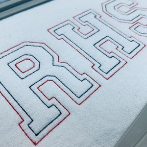 May include: Close-up of white fabric embroidered with the letters "RHS" in a collegiate style. The letters are outlined in navy blue with a red border, creating a classic, bold look. The fabric has a textured appearance.