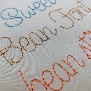 May include: A white fabric surface featuring machine embroidery in cursive script. The word Sweet is stitched in light blue, Bean Font in gold, and the partial word bean stil in orange thread, showcasing the texture of the embroidery stitches.