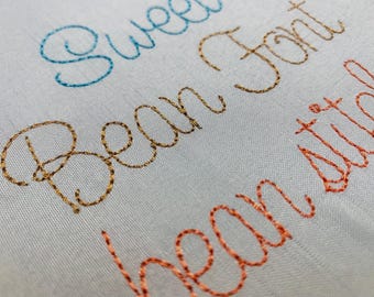 May include: A white fabric surface featuring machine embroidery in cursive script. The word Sweet is stitched in light blue, Bean Font in gold, and the partial word bean stil in orange thread, showcasing the texture of the embroidery stitches.