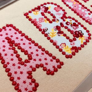 May include: An embroidery project on cream-coloured fabric featuring the letters A, B, and U. Each letter is appliqued with floral-patterned fabric in shades of pink, white, and red, and outlined with a decorative red stitched border.