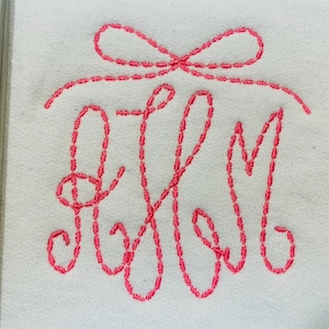 May include: A white fabric towel featuring a hot pink embroidered monogram with the letters A, H, and M in a cursive script font. Above the monogram is a decorative embroidered hot pink bow design, all stitched onto the white textile surface.