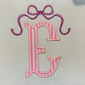 May include: An embroidered letter E on a light-coloured fabric background. The letter features a vertical pink and white striped pattern with a pink border. Above the letter is a purple embroidered bow with two long, curved ribbons extending outwards.