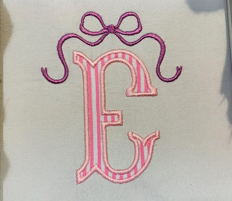 May include: An embroidered letter E on a light-coloured fabric background. The letter features a vertical pink and white striped pattern with a pink border. Above the letter is a purple embroidered bow with two long, curved ribbons extending outwards.