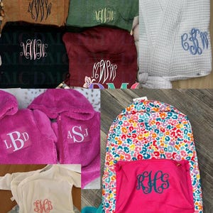 Op de afbeelding: Assortiment van gepersonaliseerde items, waaronder truien, een witte wafelbadjas, roze fleece jassen, een babyromper en een kleurrijke rugzak. Elk item heeft aangepaste geborduurde monogrammen in verschillende kleuren en stijlen.