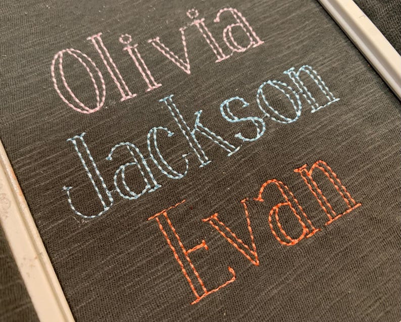 May include: A dark gray fabric surface featuring three embroidered names in a double-line stitch style. The name Olivia is stitched in light pink, Jackson in light blue, and Evan in orange, all contained within a white embroidery hoop frame.