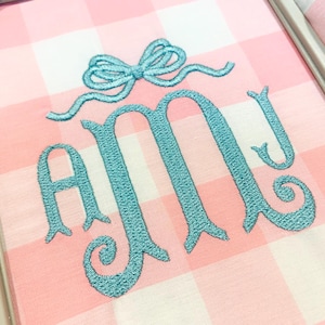 May include: A light pink and white gingham fabric piece secured in an embroidery hoop. The fabric features a teal embroidered monogram of the letters AMJ, topped with a teal embroidered bow design centred above the letters.