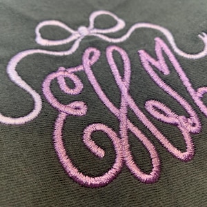 May include: A dark grey fabric featuring an embroidered monogram with the letters ELM in a cursive script. The embroidery uses a gradient thread that transitions from light lavender to a deeper purple, topped with a decorative bow design.
