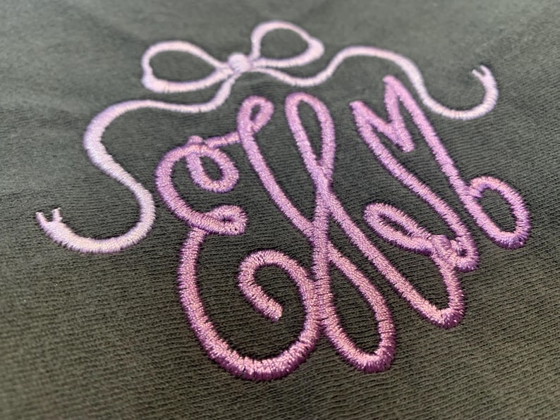 May include: A dark grey fabric featuring an embroidered monogram with the letters ELM in a cursive script. The embroidery uses a gradient thread that transitions from light lavender to a deeper purple, topped with a decorative bow design.