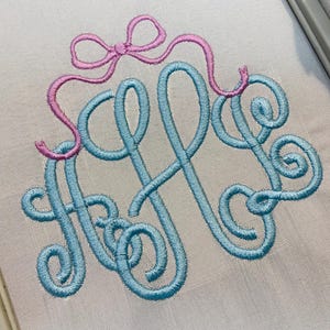 May include: Embroidered monogram in light blue thread with a pink bow accent. The monogram features elegant, swirling letters on a white fabric background. The design is likely for personalized items.