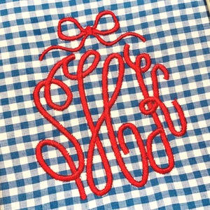 May include: A blue and white gingham fabric featuring a centered red embroidered monogram with three intertwined letters. Above the monogram is a small red embroidered bow. The fabric is secured within a circular embroidery hoop frame.