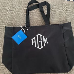 Op de afbeelding: Zwarte draagtas met witte geborduurde initialen "AGM". De tas heeft zwarte handvatten en een blauw label. De tas is gemaakt van een duurzaam materiaal en is ontworpen voor het dragen van spullen.