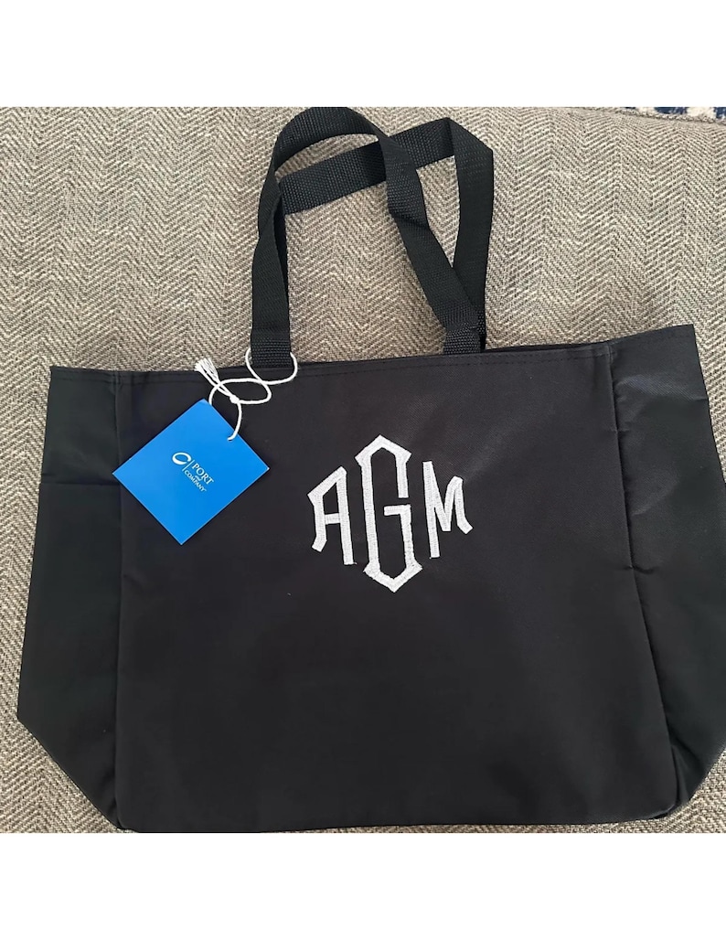 Op de afbeelding: Zwarte draagtas met witte geborduurde initialen "AGM". De tas heeft zwarte handvatten en een blauw label. De tas is gemaakt van een duurzaam materiaal en is ontworpen voor het dragen van spullen.