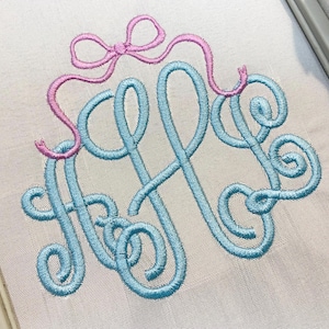 Può includere: Un monogramma ricamato con le lettere H e B in un carattere corsivo azzurro, accentuato da un fiocco rosa cucito sopra le lettere. Il disegno è ricamato su una superficie di tessuto chiaro e tinta unita all'interno di una cornice.