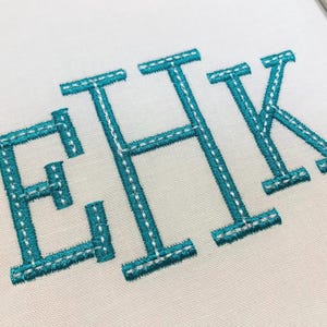 May include: The letters EHK are embroidered in a bold, teal-colored thread on a plain white fabric background. Each letter features a distinct double-stitched outline in white thread, creating a textured, block-style monogram design.