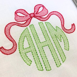 May include: A white fabric surface featuring an embroidered design of a bright red ribbon tied into a bow positioned above a large, lime green monogram consisting of the capital letters A, H, and M in a stylised, interlocking script font.