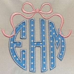 May include: An embroidered monogram featuring the letters EHM in a light blue fabric with white polka dots, outlined in a darker blue thread. Above the letters is a pink embroidered bow with long, curved ribbons extending to the sides.