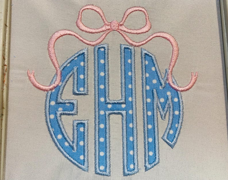 May include: An embroidered monogram featuring the letters EHM in a light blue fabric with white polka dots, outlined in a darker blue thread. Above the letters is a pink embroidered bow with long, curved ribbons extending to the sides.