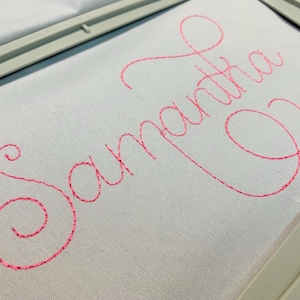 May include: Close-up of a white fabric with the name "Samantha" embroidered in a vibrant pink cursive font. The embroidery is framed by a gray hoop, suggesting a craft project or personalized item.