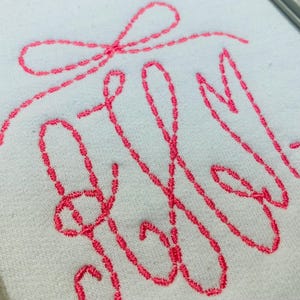 May include: A white fabric surface featuring a hot pink embroidered monogram design. The embroidery consists of three stylized, interconnected cursive letters topped with a decorative bow, all rendered in a continuous, dashed-line stitch pattern.