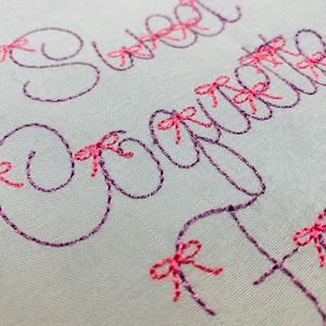 May include: A light gray fabric featuring dark purple embroidered cursive text that reads Sweet Coquette. The letters are accented with small, bright pink embroidered bows placed at various points along the stitched lettering.
