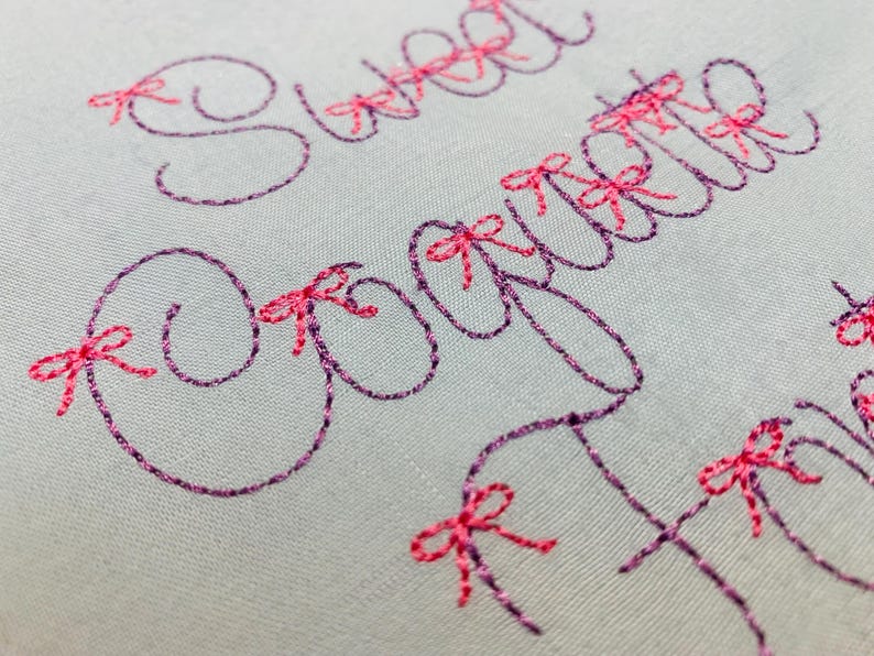 May include: A light gray fabric featuring dark purple embroidered cursive text that reads Sweet Coquette. The letters are accented with small, bright pink embroidered bows placed at various points along the stitched lettering.