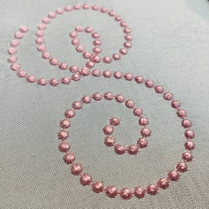 Puede incluir: Primer plano de una tela azul claro con dos diseños de espirales rosas bordadas. Las espirales están compuestas por pequeñas puntadas texturizadas y espaciadas, creando un patrón delicado y detallado. El hilo de bordar es rosa pastel.