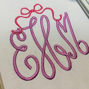 May include: A white fabric surface featuring an embroidered monogram of the letters EHM in a cursive, purple-to-pink gradient thread. Above the monogram, a small, bright pink embroidered bow is stitched with a thin, flowing ribbon detail.