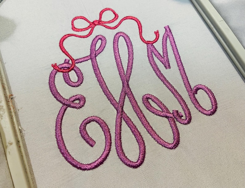 May include: A white fabric surface featuring an embroidered monogram of the letters EHM in a cursive, purple-to-pink gradient thread. Above the monogram, a small, bright pink embroidered bow is stitched with a thin, flowing ribbon detail.