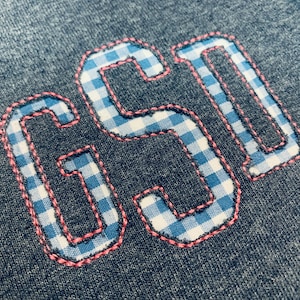 Könnte beinhalten: Nahaufnahme eines grauen Stoffes mit den gestickten Zahlen "651". Die Zahlen sind mit einem blau-weiß karierten Muster gefüllt und mit rosa Nähten umrandet. Der Stoff hat ein strukturiertes Aussehen.