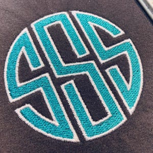 May include: A dark grey fabric surface featuring a circular monogram embroidery design. The monogram consists of three interlocking letters stitched in a vibrant turquoise thread, outlined with a thin white border for clear definition.