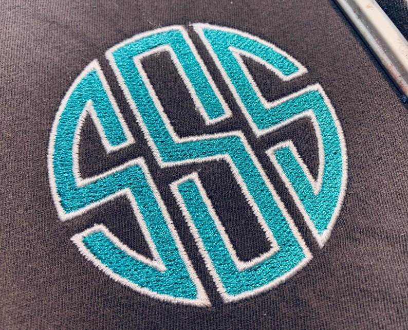 May include: A dark grey fabric surface featuring a circular monogram embroidery design. The monogram consists of three interlocking letters stitched in a vibrant turquoise thread, outlined with a thin white border for clear definition.
