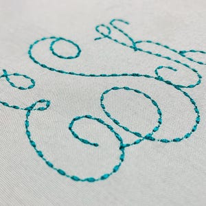 May include: A close-up of a white fabric with the word "Elle" embroidered in teal thread.