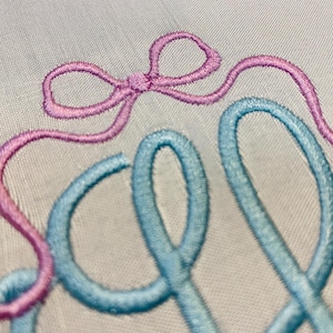 May include: Close-up of embroidered text in light blue and pink thread on a white fabric. The word "Sew" is visible, with the "S" in pink and the "ew" in light blue. A pink bow is above the "S".
