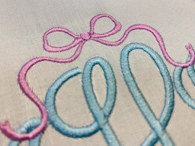 May include: Close-up of embroidered text in light blue and pink thread on a white fabric. The word "Sew" is visible, with the "S" in pink and the "ew" in light blue. A pink bow is above the "S".