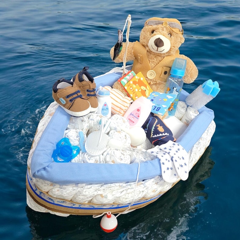 May include: A blue and white diaper cake shaped like a boat with a teddy bear wearing a fishing vest and holding a fishing rod. The boat is filled with baby items, including diapers, bottles, socks, and a pacifier.