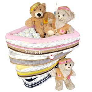 May include: A pink and white diaper cake shaped like a boat with two teddy bears wearing sailor hats. The cake is decorated with brown and white ribbon and the name "Scarlett" is embroidered on the side.
