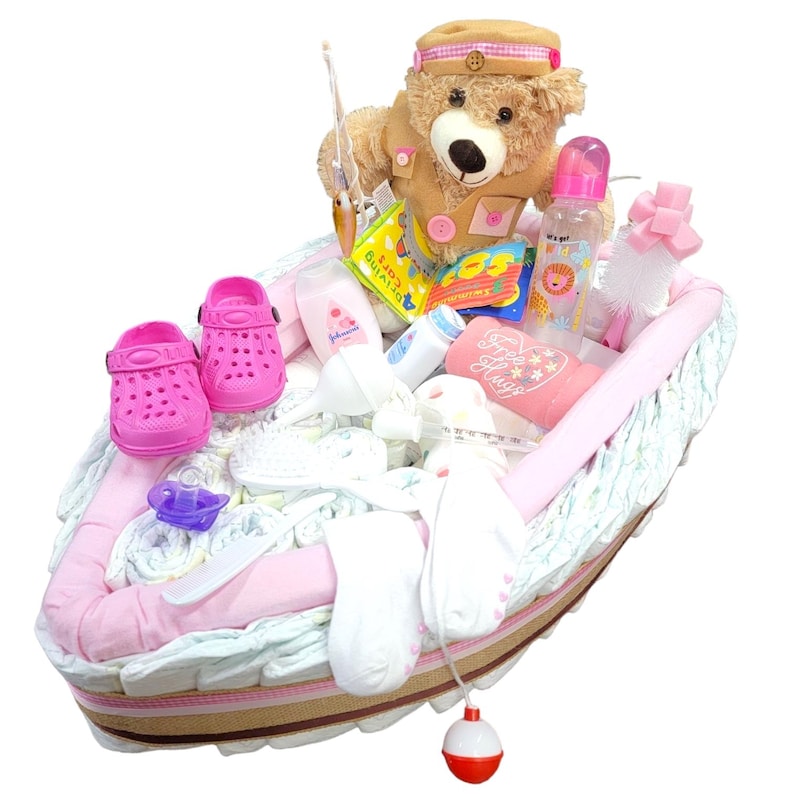 May include: A pink and white diaper cake shaped like a boat with a teddy bear wearing a fishing hat, a fishing rod, and baby items like a bottle, socks, and shoes.