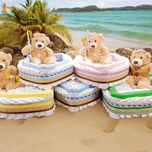 May include: Four teddy bear diaper cakes decorated as boats. Each boat is made of diapers and has a teddy bear wearing a life vest and holding a fishing pole. The boats are arranged on a sandy beach.