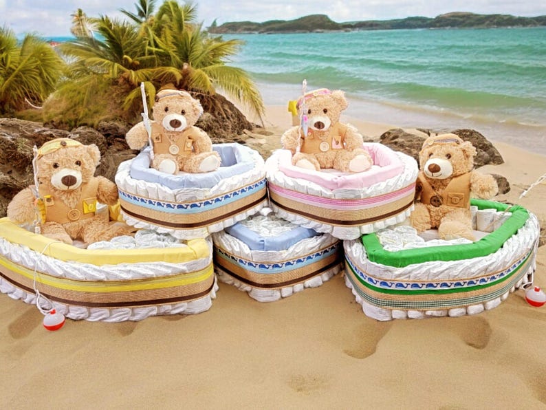 May include: Four teddy bear diaper cakes decorated as boats. Each boat is made of diapers and has a teddy bear wearing a life vest and holding a fishing pole. The boats are arranged on a sandy beach.