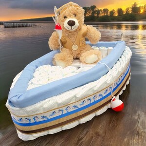 May include: A blue and brown diaper cake shaped like a boat with a teddy bear wearing a fishing hat and holding a fishing rod. The boat is decorated with blue and white dolphins and a brown ribbon.