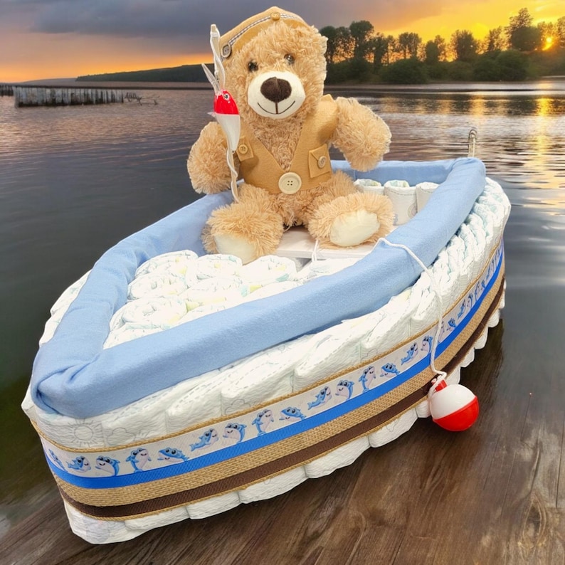 May include: A blue and brown diaper cake shaped like a boat with a teddy bear wearing a fishing hat and holding a fishing rod. The boat is decorated with blue and white dolphins and a brown ribbon.