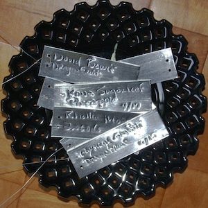 May include: A black ceramic plate with a decorative pattern of small squares.  The plate has five metal tags with handwritten text, including "David Bowie Dragon Fruit", "Kona Sugarleaf Pineapple", "Rhizoma Mosa", "Physical Graffiti", and "Despereaux Flax".