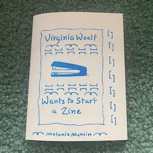 May include: A white zine with blue ink text and illustrations. The cover reads Virginia Woolf Wants to Start a Zine by Melanie Maksin. It features a central drawing of a stapler, decorative border lines, and a vertical column of bracket symbols.
