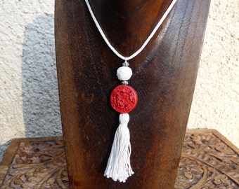 Puede incluir: Un collar de cordón blanco con un colgante rojo y blanco y una borla blanca. El colgante es un botón redondo rojo con un diseño blanco.