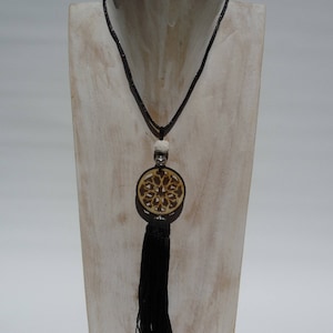 Peut inclure: Un long collier noir avec un pendentif circulaire et une longue gland. Le pendentif est de couleur marron clair avec un motif découpé. Le collier est présenté sur un présentoir à bijoux en bois blanc.
