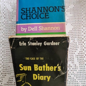 May include: Two paperback books stacked on top of each other. The top book is blue with white text that reads "Shannon's Choice" by Dell Shannon. The bottom book is black with yellow text that reads "The Case of the Sun Bather's Diary" by Erle Stanley Gardner. A Perry Mason Mystery.