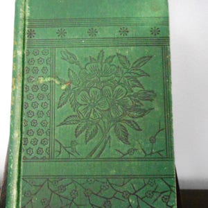 May include: A green book with a floral design on the cover. The design features a bouquet of flowers with leaves and stems. The flowers are stylized and have a dark green color. The book has a decorative border around the edges.