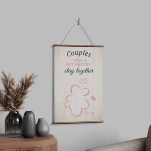 Op de afbeelding: Een witte canvas wandhanger met een roze hartontwerp en de tekst "Couples that fart together, stay together".