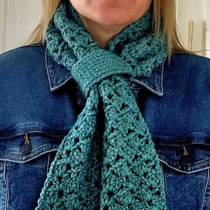 May include: A teal crocheted scarf with a delicate openwork pattern. The scarf is tied around the neck of a person wearing a denim jacket.