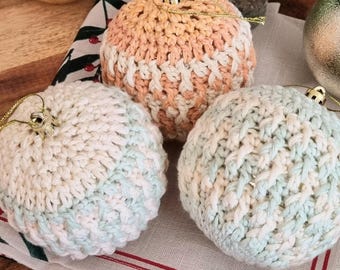 May include: Three crocheted Christmas ornaments in different colors. The ornament on the left is white and light green, the ornament in the center is peach and white, and the ornament on the right is white and light blue.